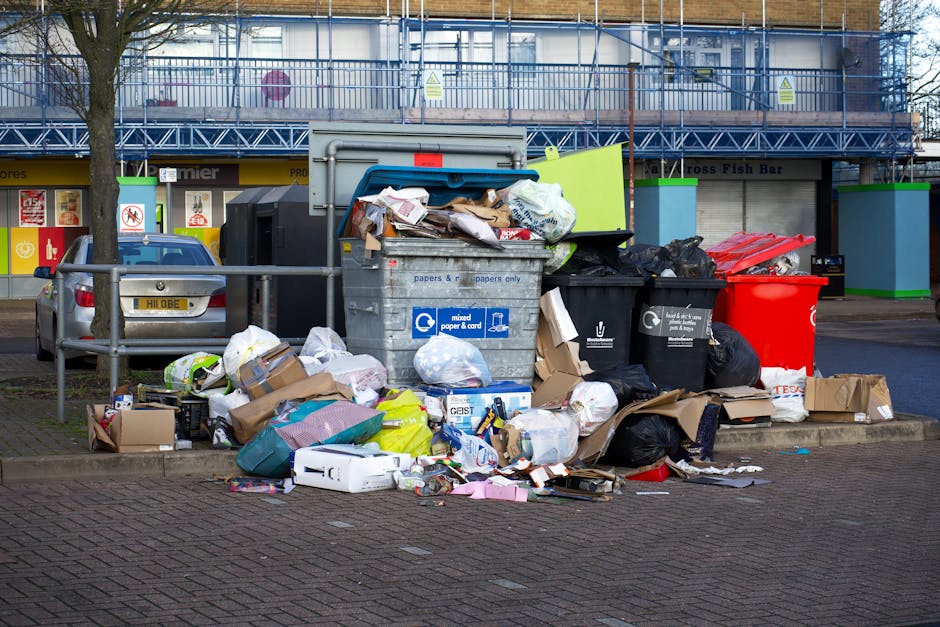 A row of large, rectangular waste bins positioned along a paved sidewalk next to a brick wall, each with a different coloured lid for segregated rubbish collection, including green, blue, red, and dar
