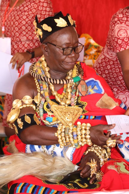 A woman dressed in traditional African attire is seated, wearing an elaborate headdress decorated with gold accents and a crown-like structure. She is adorned with multiple layers of large, ornate nec